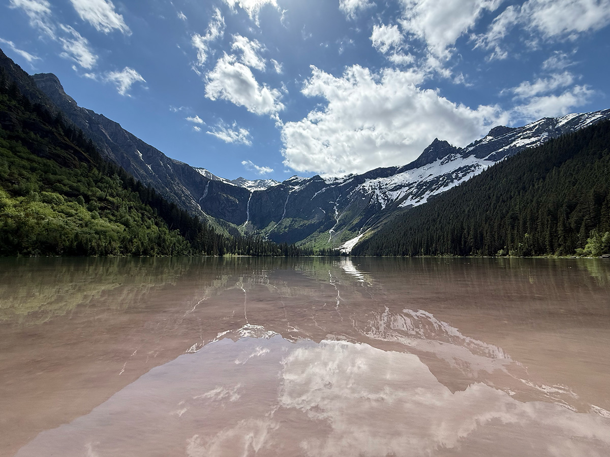 Avalanche Lake in Glacier National Park, still water reflecting snow-streaked peaks and forested slopes under a bright sky.