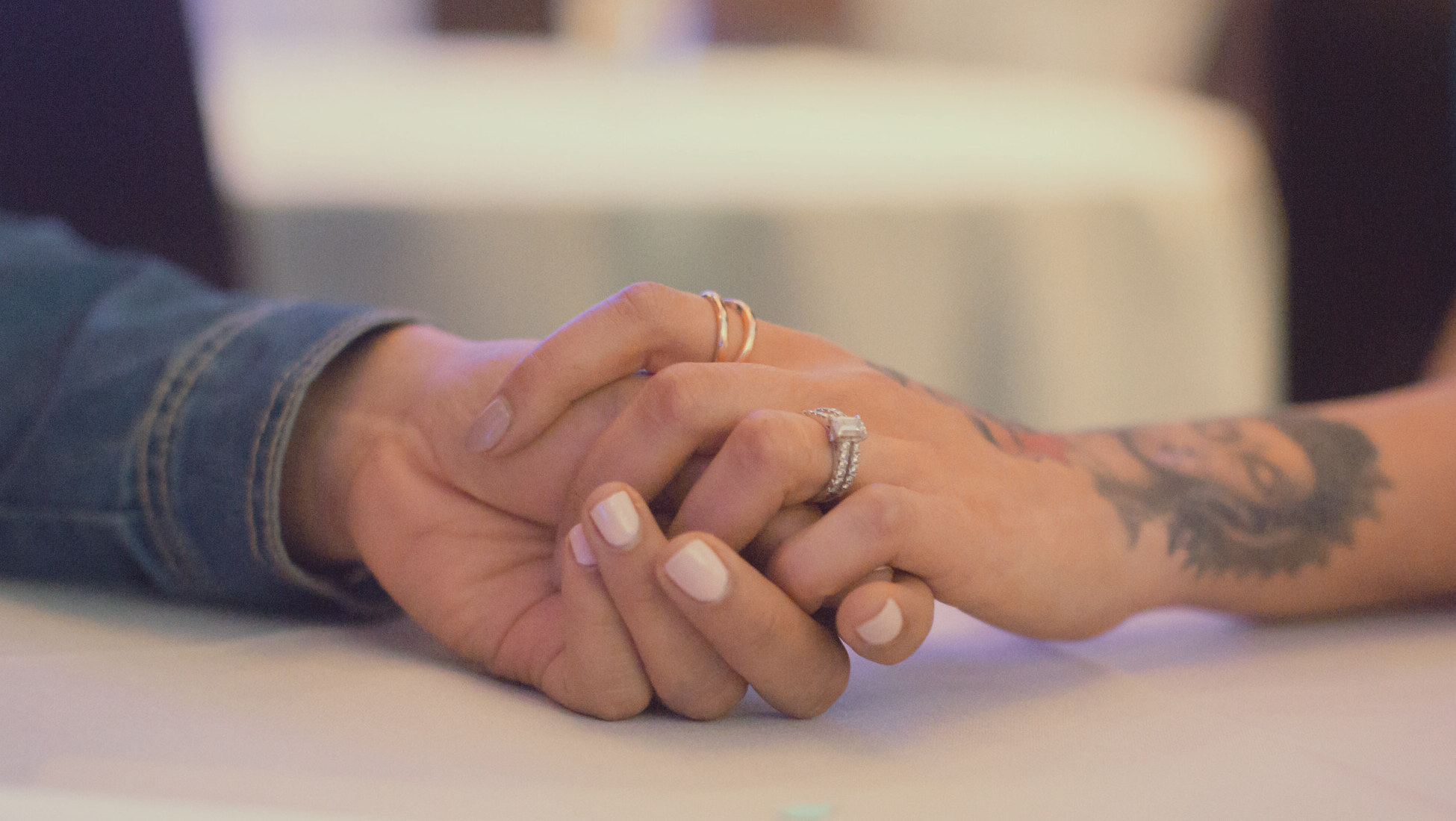 Two people's hands resting together on a white table, fingers loosely interlaced — one wrist in a denim cuff, the other forearm tattooed.