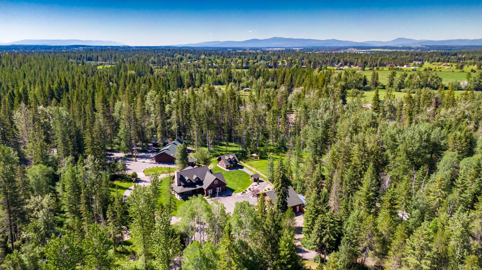 Blue Star Resort and Retreat from the air: dark-timbered lodges and cabins gathered around a clearing deep in Montana pine forest, with open pasture and a line of blue mountains beyond.