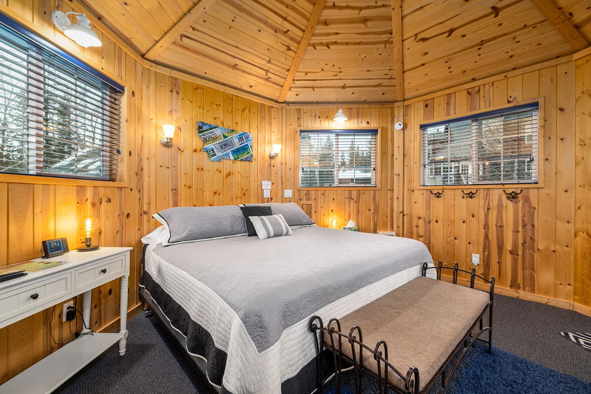 Inside the bungalow: a knotty-pine room with a panelled octagonal ceiling, a king bed made up in grey and white, a bench at its foot, a writing desk under a window, and pines through the blinds on three sides.