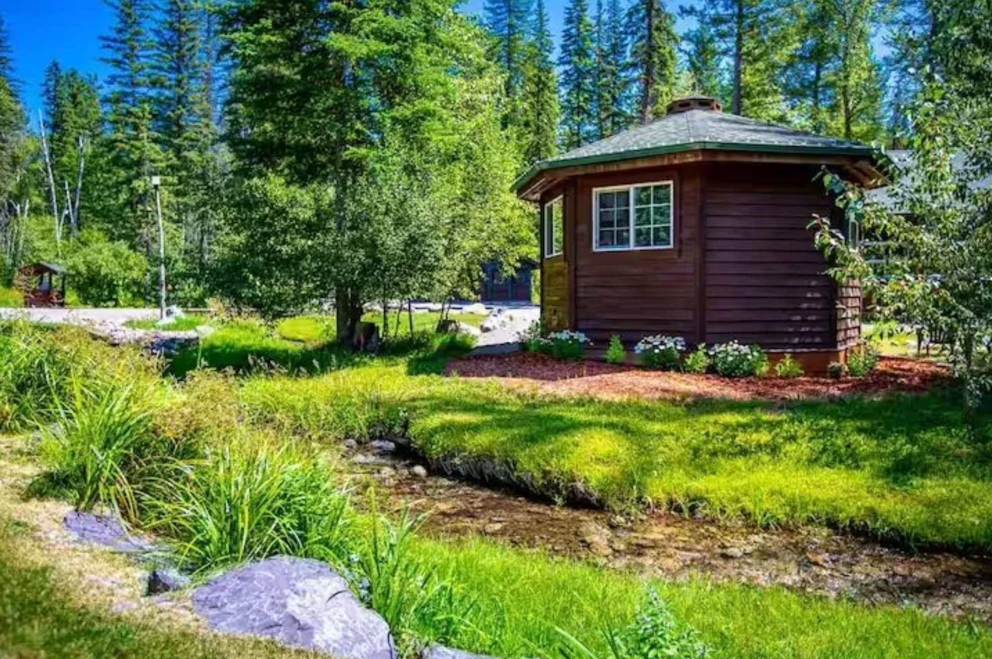 The bungalow from outside: an octagonal timber cabin with a shingled roof and a white-framed window, standing in mown grass beside a shallow creek with pines behind it.