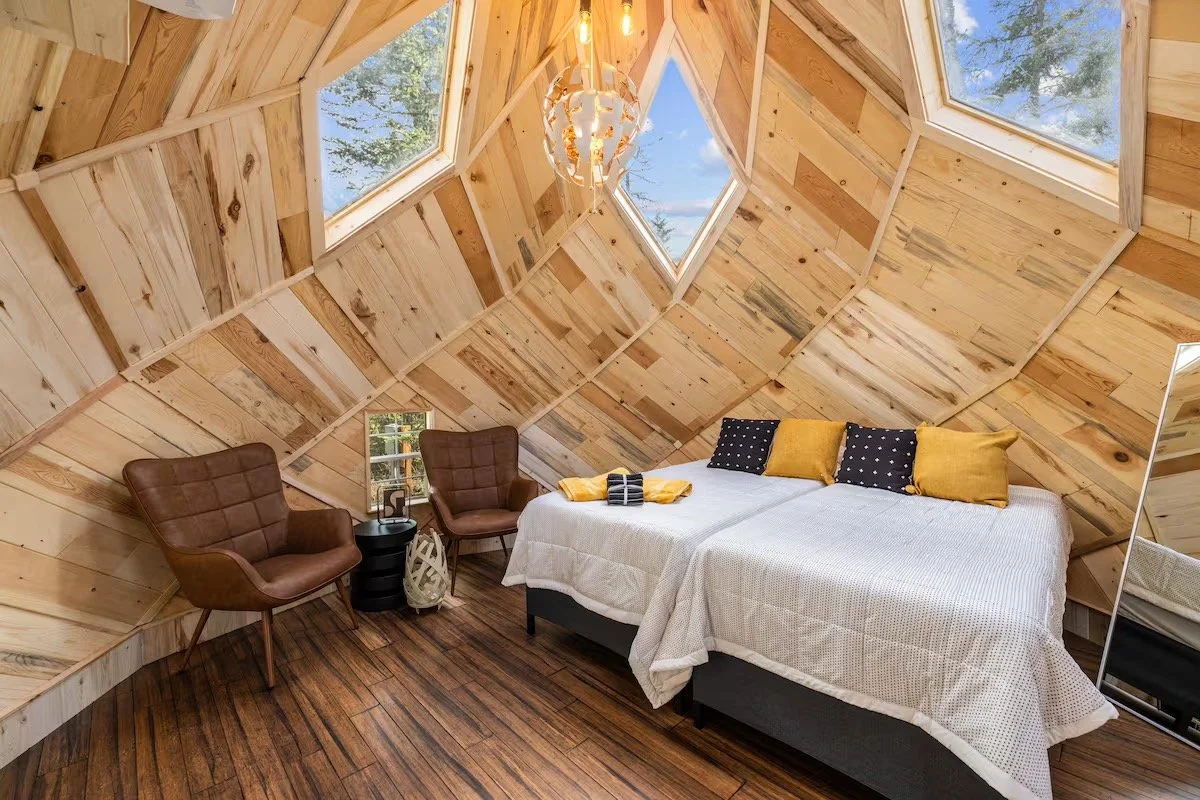 Inside a dome: pale timber panelling on every facet, a made bed with mustard and navy cushions, two leather armchairs by a small window, and a pendant light overhead.