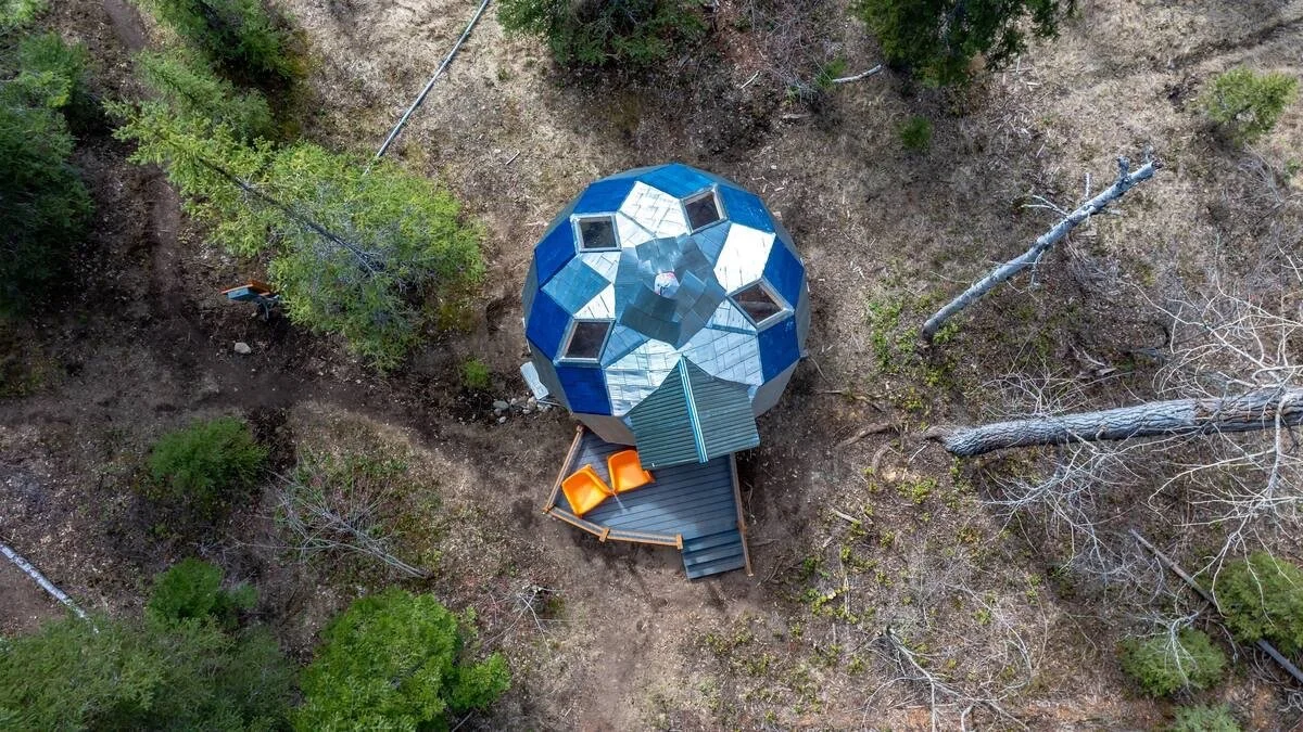A geodesic dome seen from directly overhead, its blue and mirrored panels set among pines, with a small timber deck and two orange chairs at its entrance.