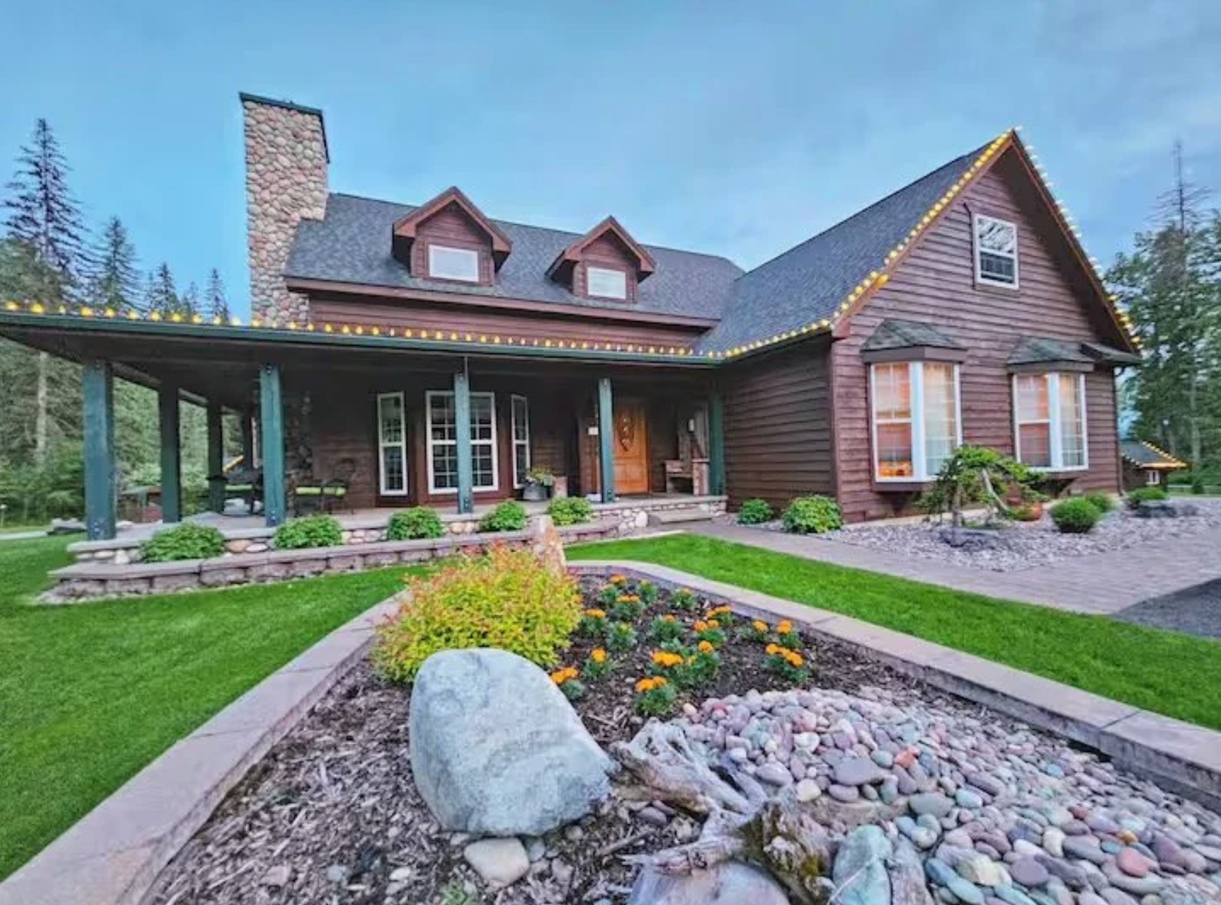 The main lodge from the front at dusk: a two-storey timber house with a stone chimney, a deep covered porch and dormer windows, its eaves strung with warm lights above clipped lawn and flower beds.