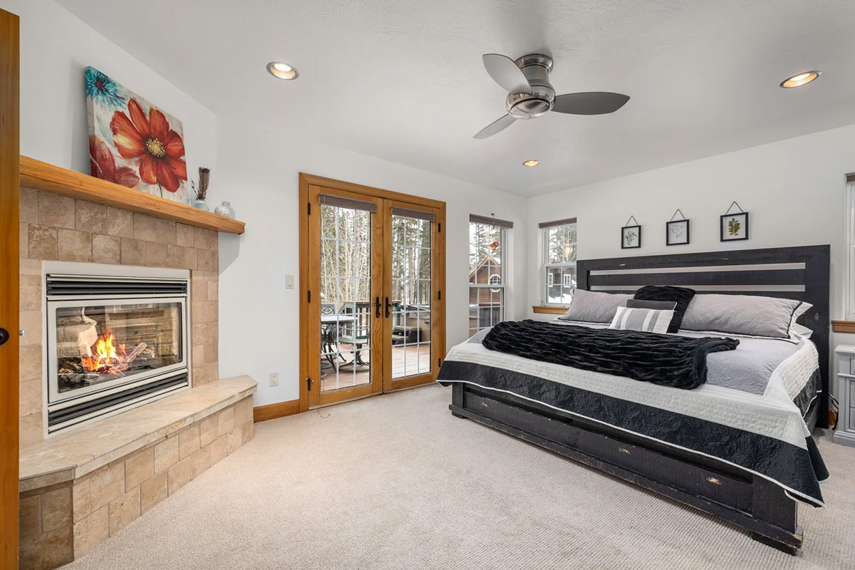 The main lodge&rsquo;s master suite: a bright white room with a lit stone fireplace, a king bed against a dark padded headboard, and French doors opening onto a deck in the trees.