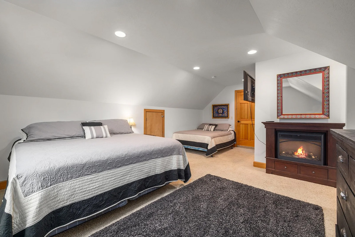 The main lodge&rsquo;s shared room, tucked under a sloping ceiling: a king bed and a second smaller bed against the far wall, a lit fireplace between them, and a wide grey rug on pale carpet.
