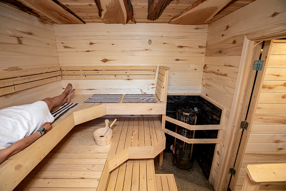 Inside the cedar sauna: tiered benches running around two walls, a stove of dark stones behind a timber guard, a wooden ladle resting in its bucket on the floor, and someone lying along the lower bench wrapped in a white towel.
