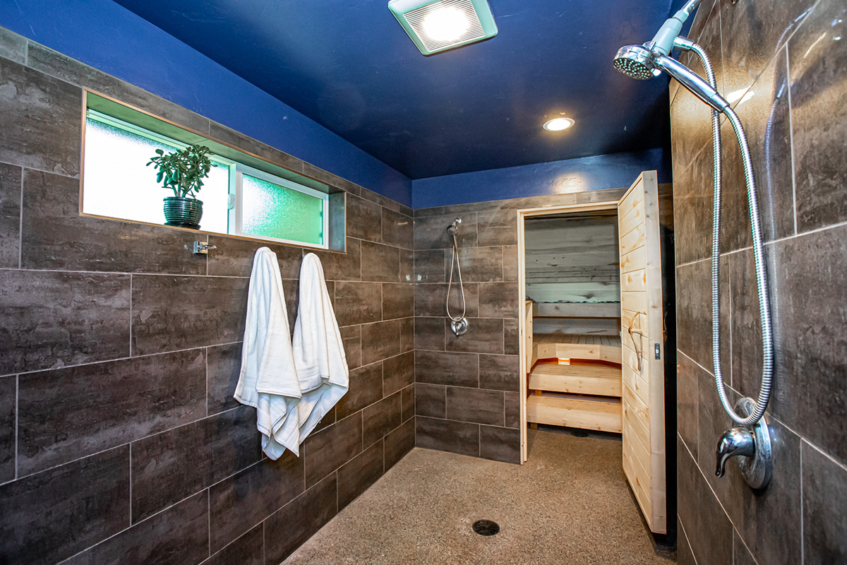The shower room outside the sauna: dark tiled walls under a deep blue ceiling, two white towels on hooks, an open rain shower, and the sauna&rsquo;s timber door standing open onto its benches.