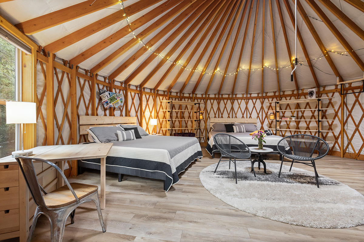 Inside a yurt: two made beds against a lattice wall of pale timber, the canvas roof rising to a ring of rafters strung with fairy lights, and two round chairs with a small table on a rug in the middle of the floor.
