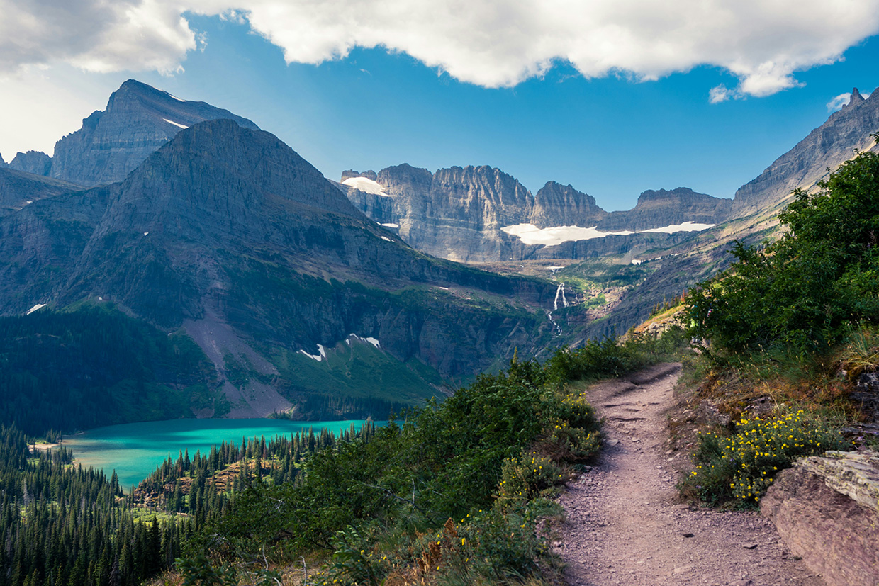A narrow dirt trail in Glacier National Park running along a slope of wildflowers toward a headwall of grey peaks streaked with snowfields, a turquoise glacial lake in the valley below.