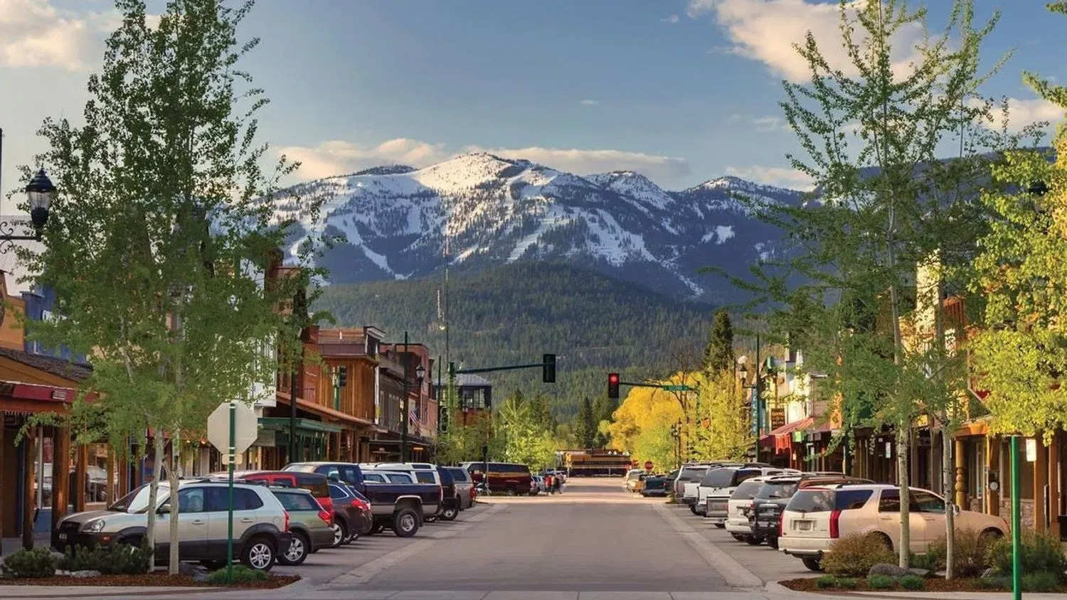 Downtown Whitefish in early evening: a main street of low timber and brick storefronts lined with parked cars and young aspens, and the snow-streaked bulk of Big Mountain filling the view at the end of it.