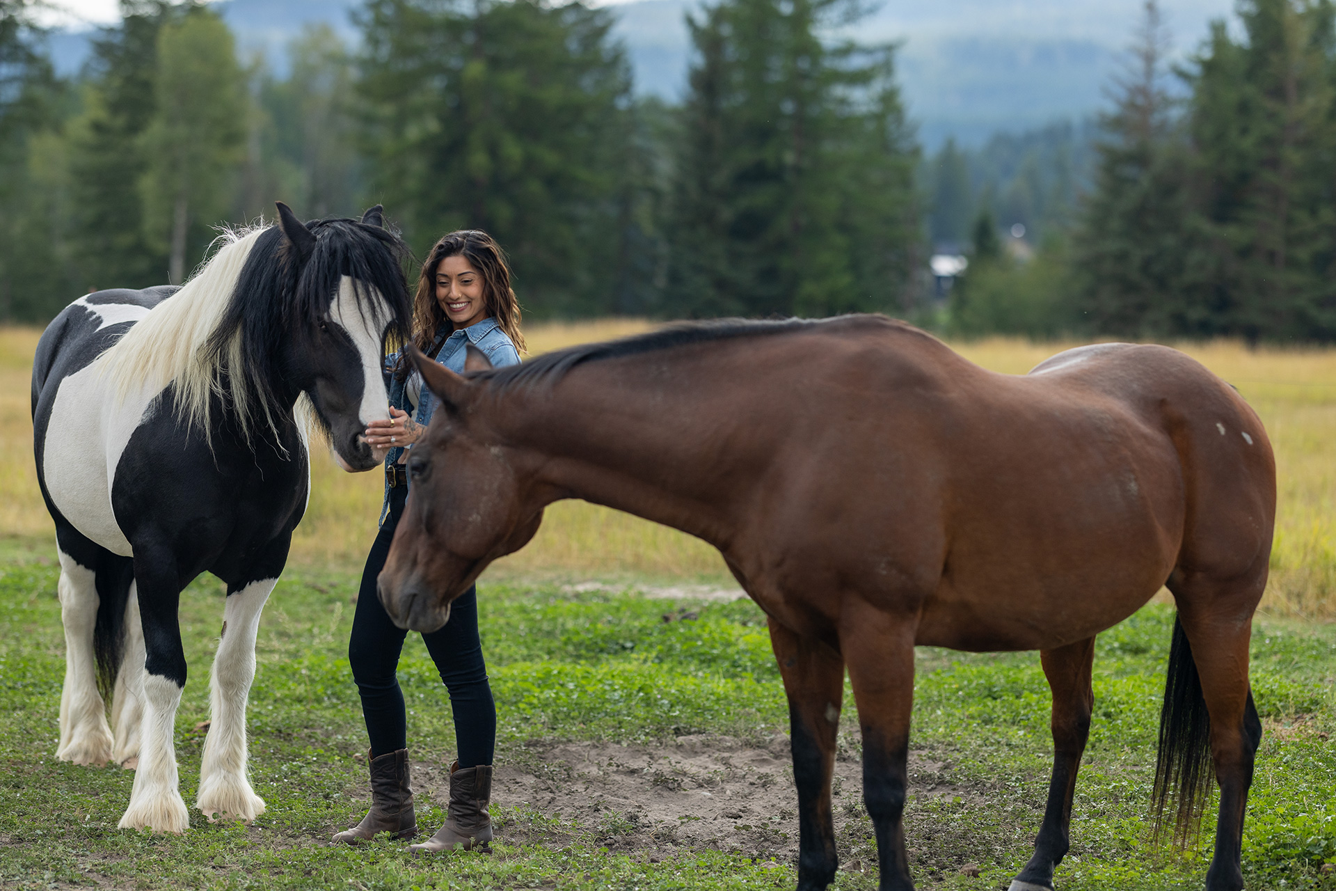 Ruby Fremon standing between two horses in a paddock, reaching up to greet a black-and-white paint while a bay grazes across the foreground.