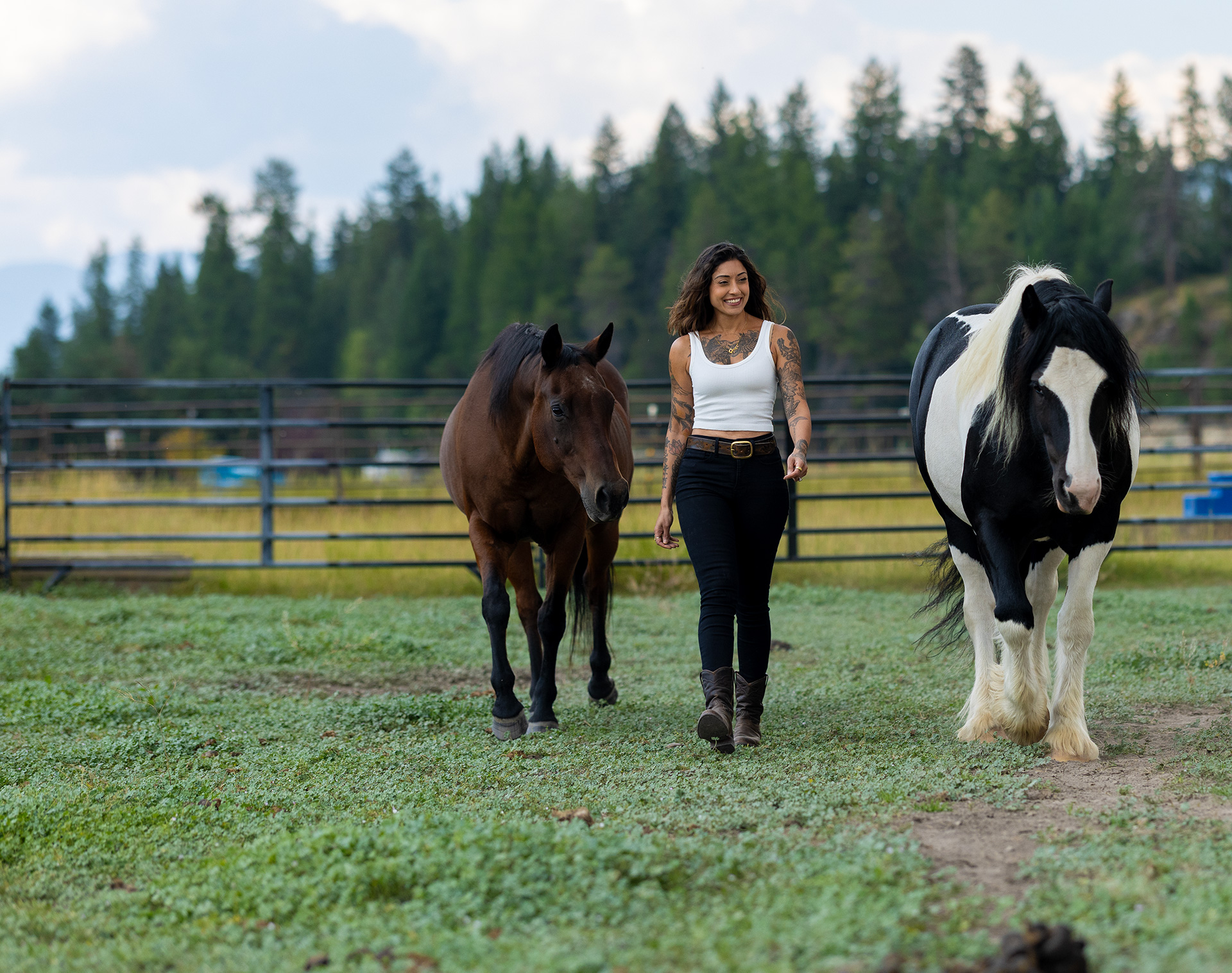 Ruby Fremon walking toward the camera across a green paddock, a bay horse loose at her left shoulder and a black-and-white paint at her right, a fence line and pine forest behind them.