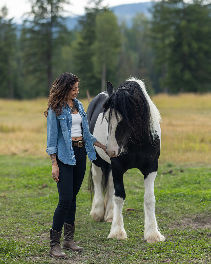 Ruby Fremon in a denim shirt standing in a Montana pasture beside a black-and-white paint horse, her hand at its muzzle and her head turned toward it, pines and a far ridge behind them.