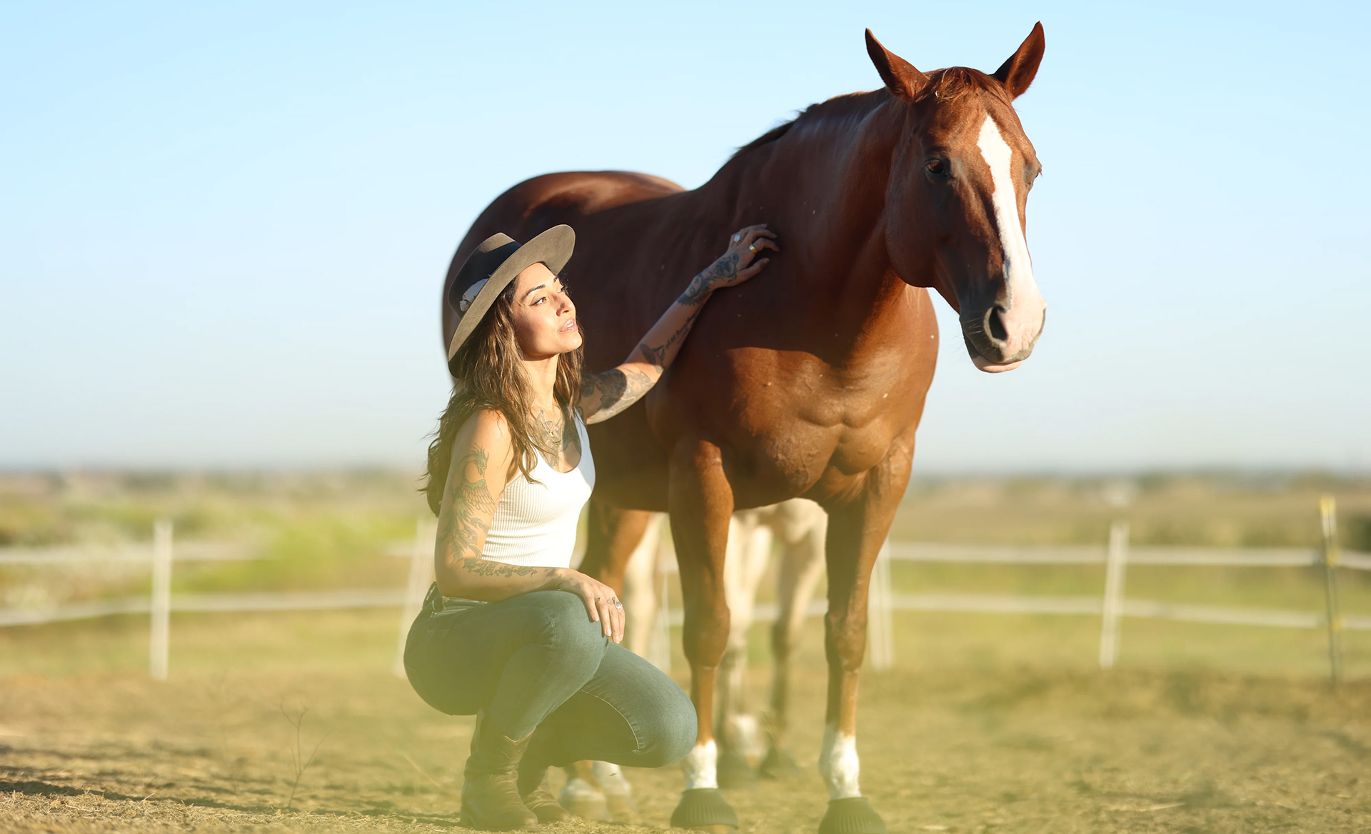 Ruby Fremon crouching in an open paddock beside a chestnut horse, both at rest under a clear sky.