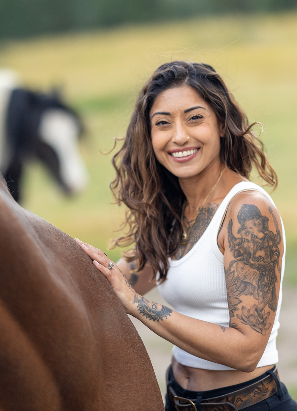Ruby Fremon standing at a bay horse's shoulder with one hand resting on its back, turning to the camera and smiling, a paint horse grazing behind her.
